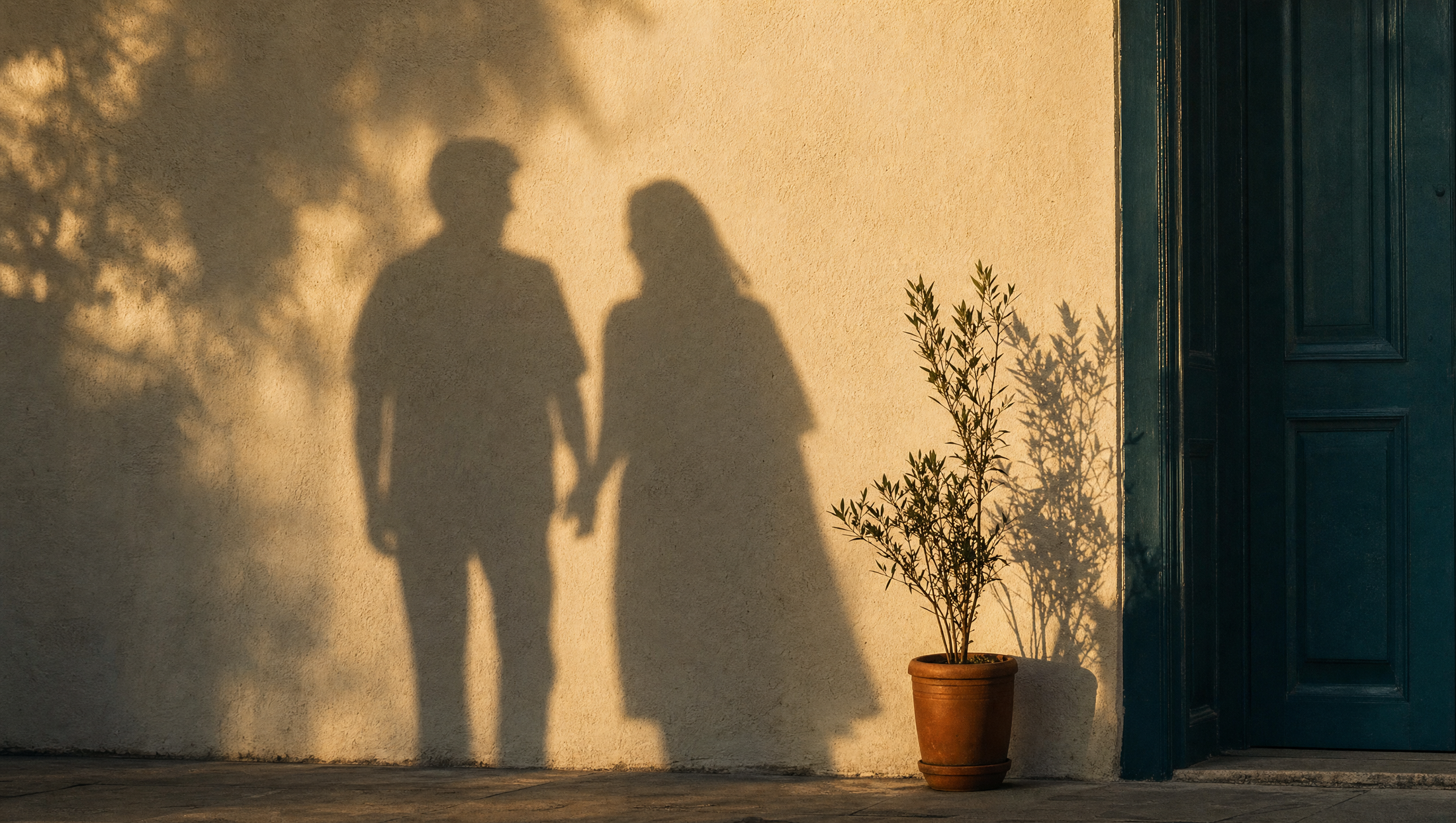 The long shadows of two people standing close together on a warm cream wall at golden hour, with a deep teal door at the edge of the frame.